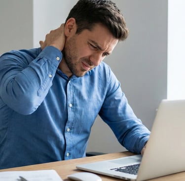 man sits at a desk with a laptop, rubbing the back of his neck in discomfort, depicting a tension-type headache