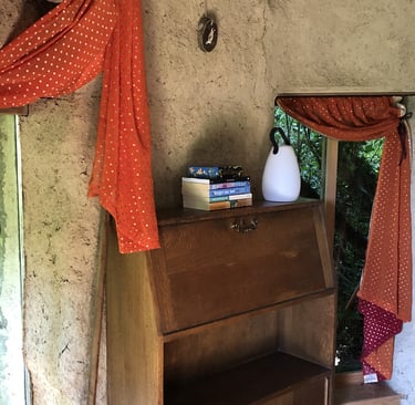 interior of a cob round-house showing a bureau, orange curtains and a pile of books