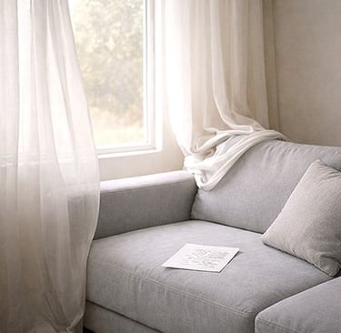 A softly lit couch by a window with sheer curtains and a single sheet of paper resting on the seat.