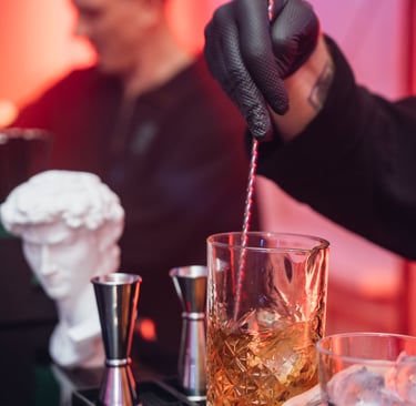 Stilingai apšviestame renginyje barmenas maišo kokteilį stiklinėje, naudojant baro šaukštą