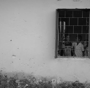 Black and white photo of three smiling children looking out of a metal barred window in a white wall.
