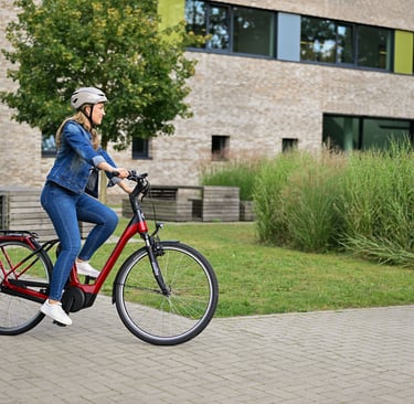 A woman wearing a helmet rides a red electric commuter bike on a paved path near a modern building.