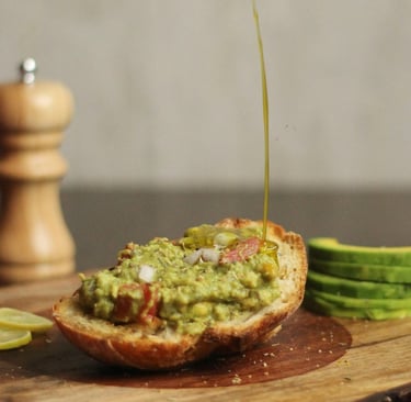 Fresh avocado toast on rustic wood being drizzled with olive oil next to a pepper grinder and lime slices.