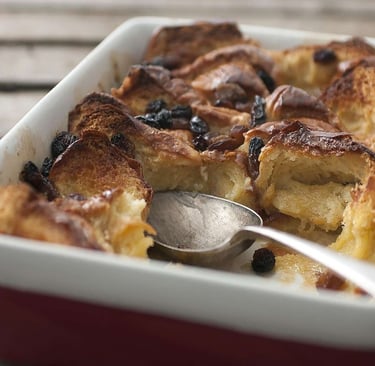 Golden brown bread and butter pudding with raisins in a ceramic baking dish with a serving spoon.