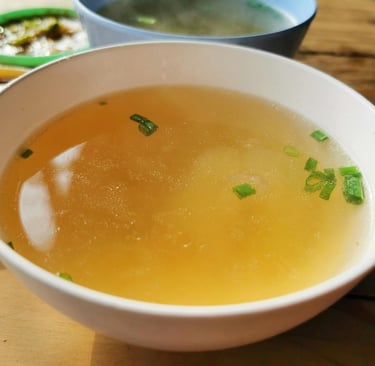A white ceramic bowl of clear chicken broth garnished with chopped green onions on a rustic wooden table.