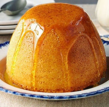 A classic golden syrup steamed sponge pudding on a blue rimmed plate with syrup drizzled over the top.
