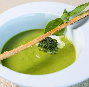 Creamy green broccoli soup in a white bowl garnished with a breadstick and fresh herbs.