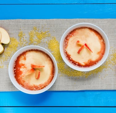 Two bowls of hot millet porridge with apple slices and sauce on a blue table.