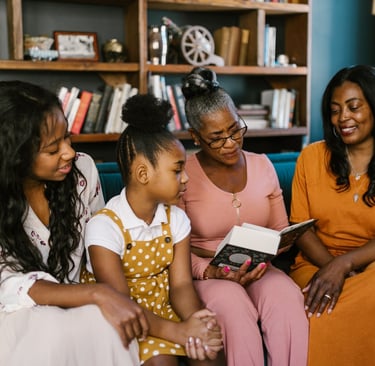 Three generations of Black women sitting on a sofa reading a book together in a library.