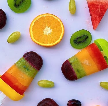 Layered fruit juice popsicles surrounded by fresh orange, kiwi, and watermelon slices on a white surface.