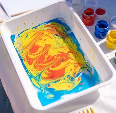 A white tray of colorful marbled paint swirled in yellow, red, and blue for ebru art.
