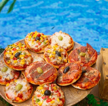 Mini pizzas with assorted toppings on a wooden platter by a blue swimming pool.