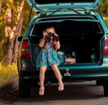 A young girl sits in a car trunk using binoculars during a family road trip adventure.