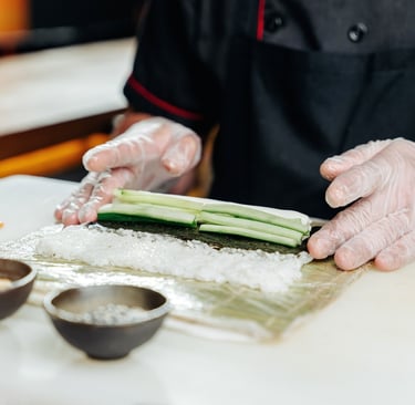 Professional chef wearing gloves preparing a fresh sushi roll with cucumber, rice, and nori seaweed.