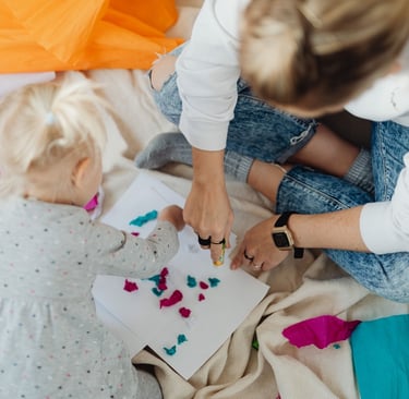 Mother and toddler girl creating colorful paper crafts with glue and tissue paper on a blanket.