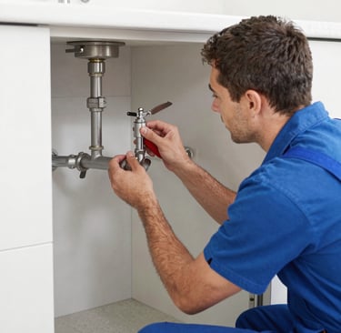 A cleaning cart is positioned in an outdoor area with a concrete building structure in the background. The cart holds a large bucket and cleaning supplies, including a striped cloth and a pink cloth hanging over the handle. A green scrubbing brush is placed on the bottom platform. Surrounding the area are vibrant flower beds with yellow and white flowers, as well as bushes and trees, contributing to a tidy and well-maintained environment.