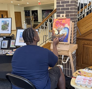 An artist with braided hair painting a colorful landscape canvas on an easel at an indoor art gallery.