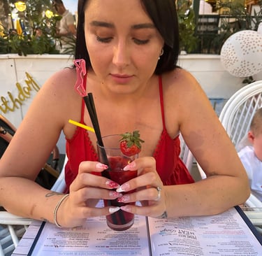 Young lady in red dress on vacation, holding a cocktail in a restaurant brilliantly displaying beautiful nails by Little Lift