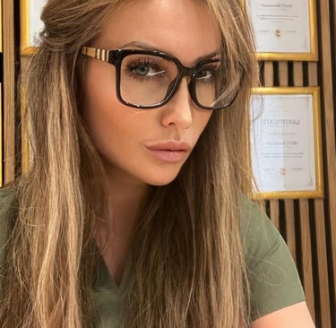 Woman with long blonde hair wearing stylish black square-frame glasses in front of framed certificates.