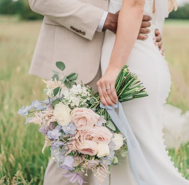 a bride and groom standing in a field, the bride is holding a pastel flower wedding bouquet