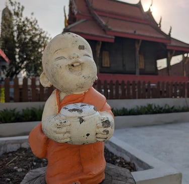 A smiling monk statue in orange robes at a Buddhist temple in Thailand during sunset.
