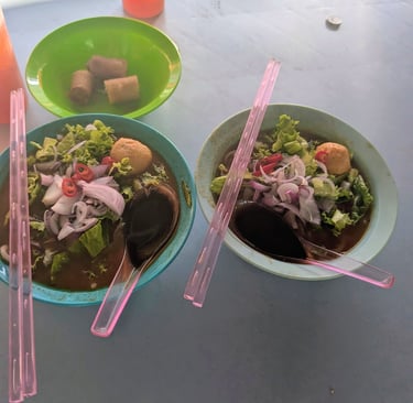 Two bowls of spicy Malaysian laksa soup with noodles, onions, and pink chopsticks on a table.