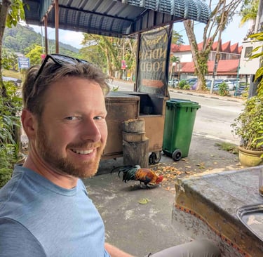 The blog's author Don sitting near a rooster in a restaurant in Penang, Malaysia.