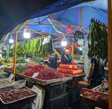 Night market stall in Malaysia selling fresh red chilies, stink beans, and local produce under a blue tent.