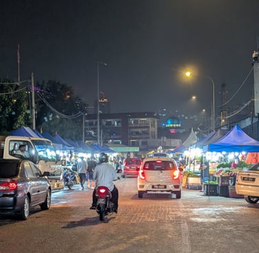 Busy night market street in Malaysia with cars, motorcycles, and blue food stalls under streetlights.