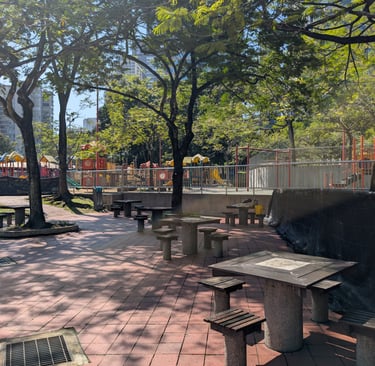 Sunny KLCC park with stone picnic tables and benches near a children's playground and green trees.