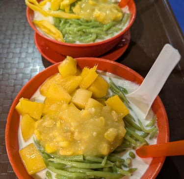 Two bowls of Malaysian Cendol dessert with green pandan noodle, fresh mango, corn, and coconut milk.