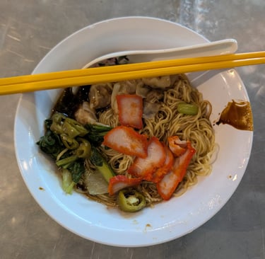 A bowl of wonton noodles with char siu pork, leafy greens, and yellow chopsticks on a metal table.