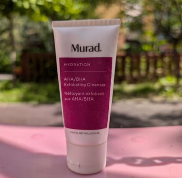 Murad Hydration AHA BHA Exfoliating Cleanser tube on a pink surface with a sunny outdoor background.