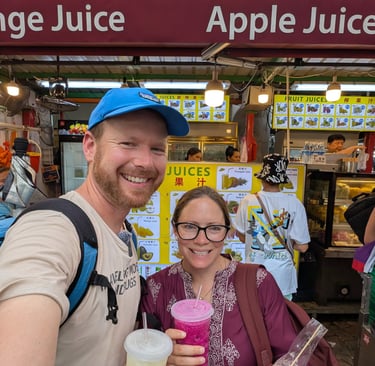 Don and Samantha holding fresh fruit juices and snacks at a vibrant Asian street food market stall.