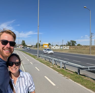 Don and samantha next to a highway road sign that sayd Novi Sad in Cyrillic 