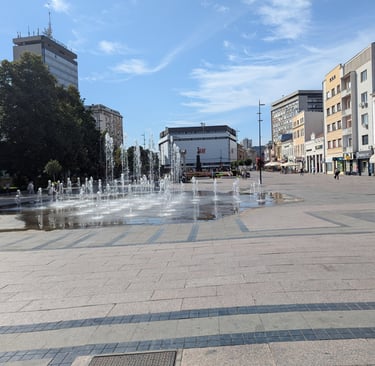 A wide stone plaza featuring a floor level water fountain and surrounding urban city buildings in NIs