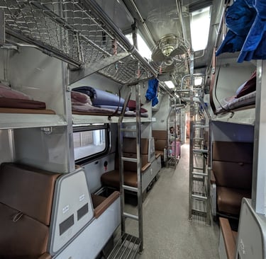 Interior of a Thai railway sleeper class coach featuring bunk beds, luggage racks, and ceiling fans.
