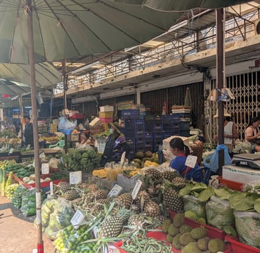 street market in Thailand selling fresh pineapples, bananas, and tropical vegetables under green umbrellas.