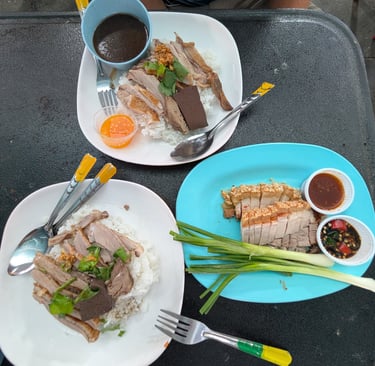 Plates of authentic Thai roasted duck over rice and crispy pork belly served with dipping sauces on a table.