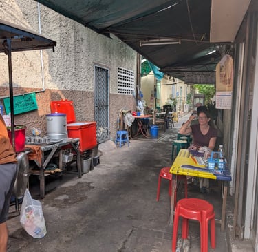 A local street food stall in a narrow Bangkok alley with plastic stools and outdoor seating.