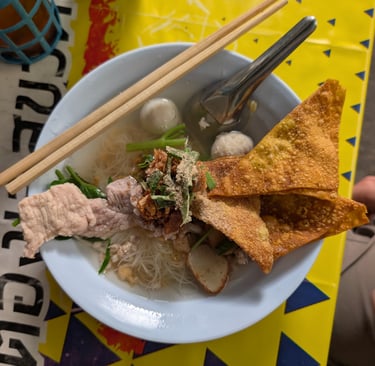 A bowl of Thai noodle soup with crispy wontons, fish balls, and pork on a yellow patterned table.