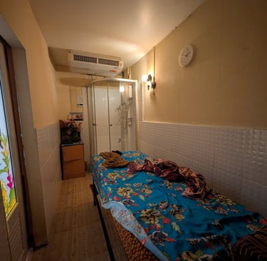 A narrow massage room with a floral blue bed, glass shower cubicle, and air conditioning unit.