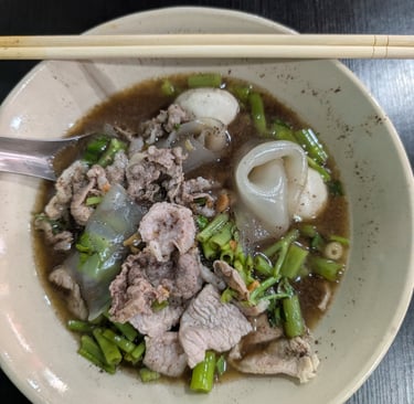 A bowl of Thai boat noodles with sliced pork, fish balls, and morning glory in a rich savory broth.