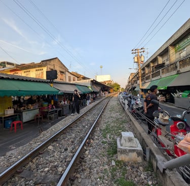 A narrow railway track runs through the Tala Market in Thailand with shops and stalls on both sides.