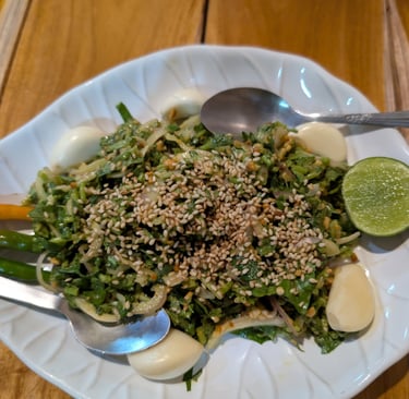 A fresh Burmese pe salad topped with toasted sesame seeds, garlic cloves, and lime on a white plate.