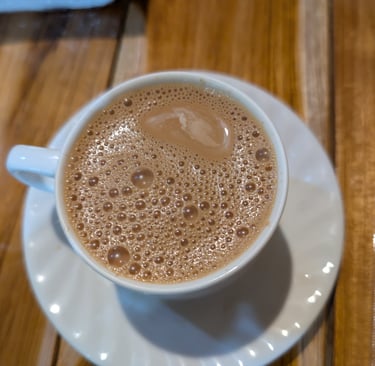 A cup of hot frothy Burmese sweet tea on a white saucer on a wooden table.
