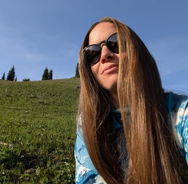 A long-haired woman wearing sunglasses enjoys a sunny day on a grassy green hillside under a clear blue sky.