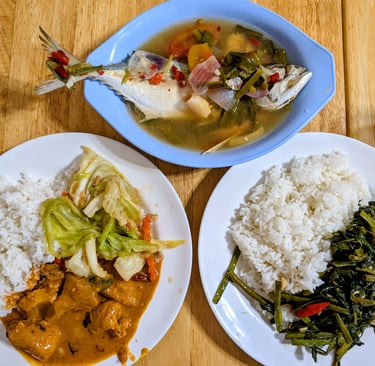 Thai steamed fish soup with white rice, red curry chicken, and stir-fried morning glory on a wooden table.