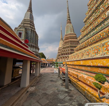 Wat Pho temple complex in Bangkok, Thailand, featuring intricate stupas with colorful ceramic decorations under a cloudy sky.