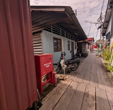 A narrow wooden boardwalk pathway in a Malaysian Yeoh Jetty with stilt houses and parked motorbik